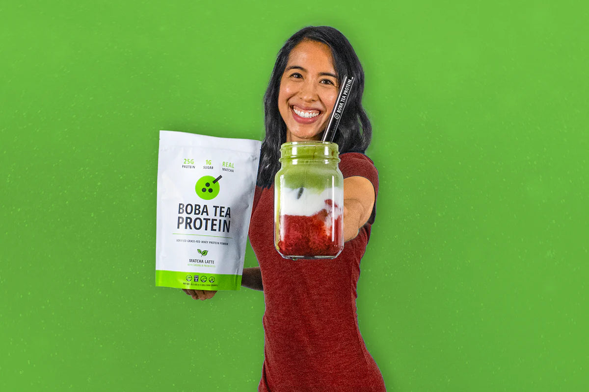 Woman holding Matcha Latte Boba Tea Protein pack and a layered matcha drink in front of green background