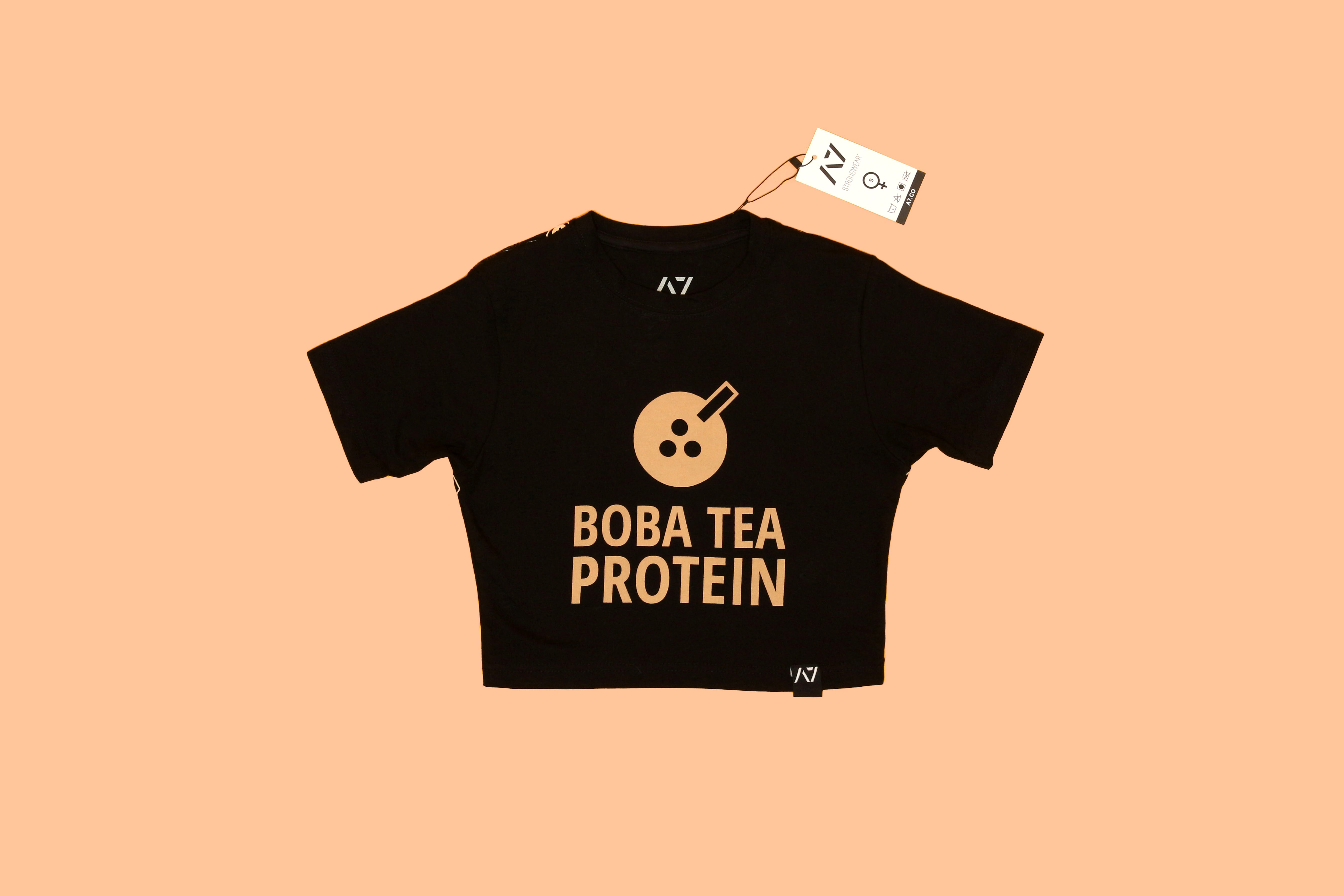 Black crop top with large "BOBA TEA PROTEIN" text and icon, displayed on a peach-colored background with a tag.