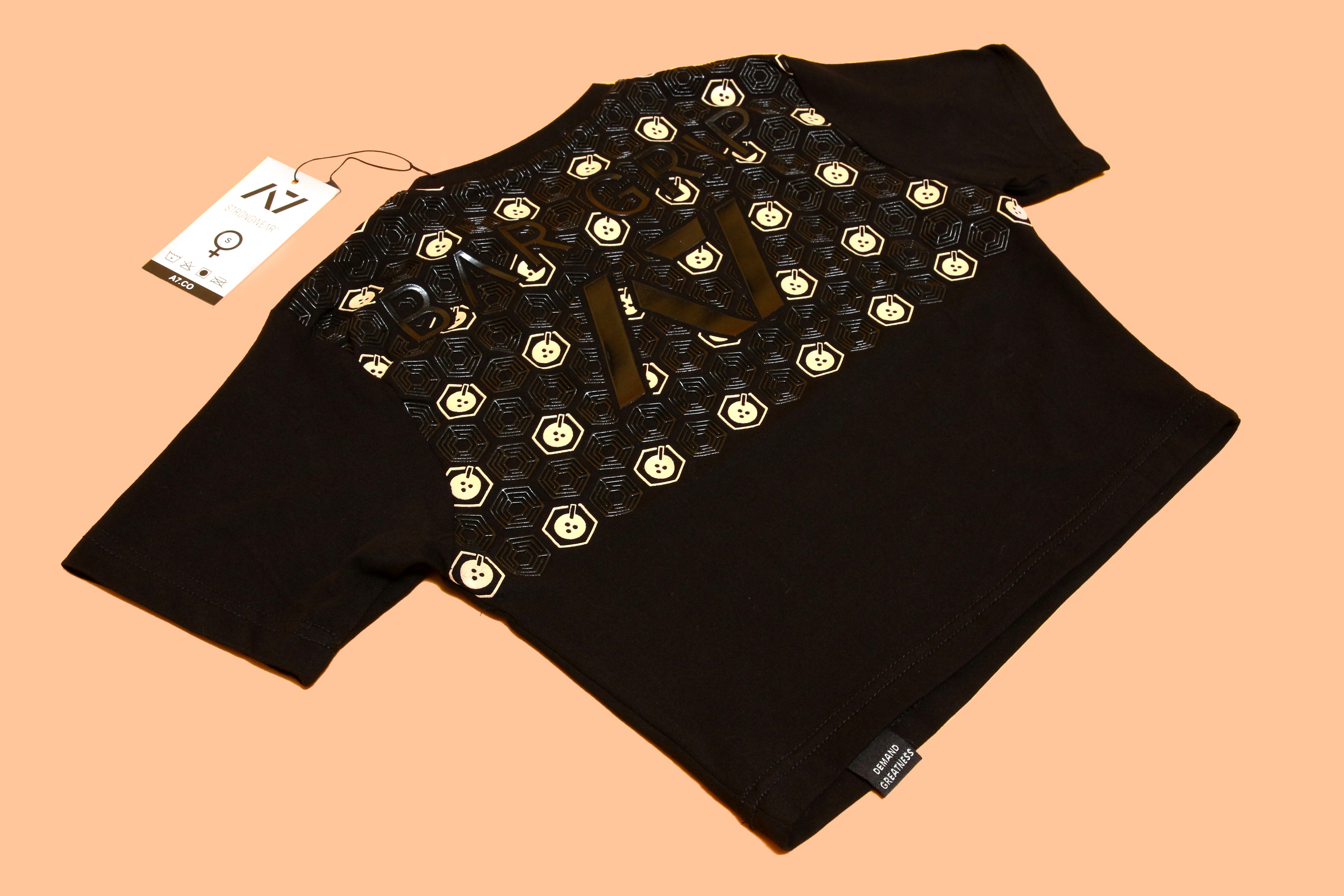 Black crop top with honeycomb and smiley face pattern, featuring bold text and a clothing tag on a peach background.