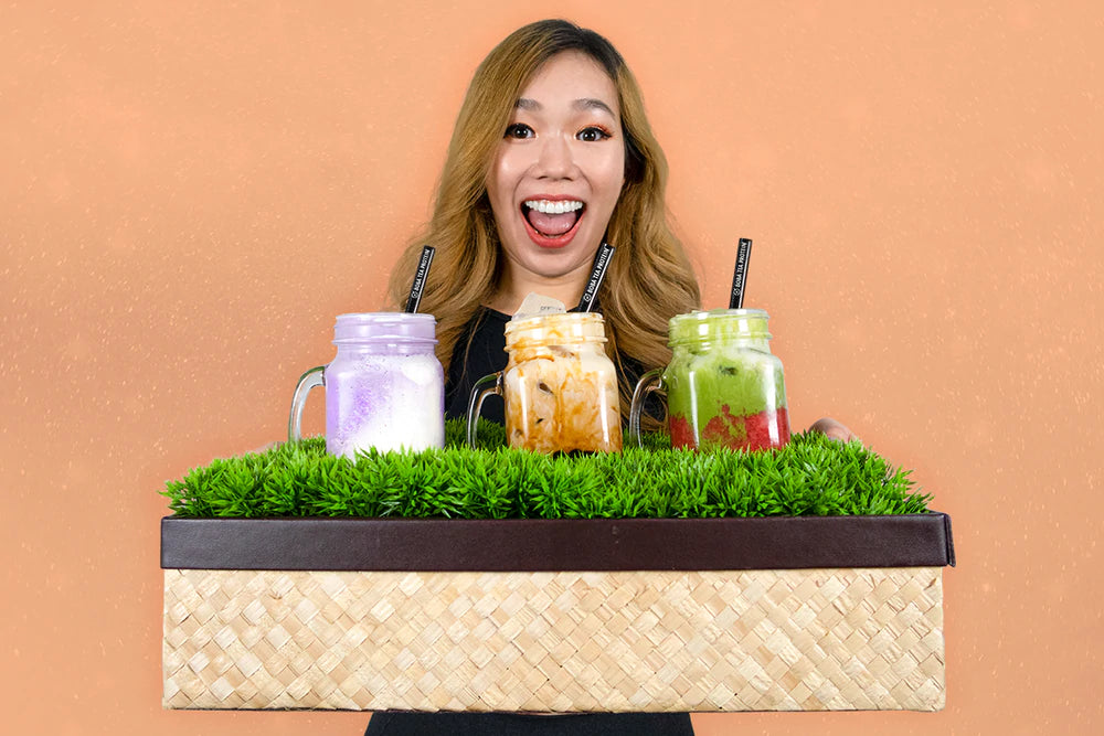 Smiling woman holding a tray with three boba tea drinks in jars, featuring taro, classic, and matcha flavors.
