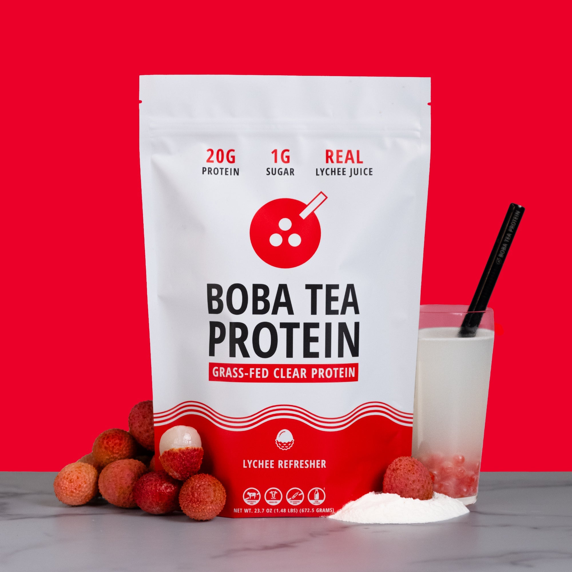 A stand-up pouch of Boba Tea Protein grass-fed clear protein in Lychee Refresher flavor, shown on a gray surface with lychee fruits, a glass of white drink with red popping pearls, and a black straw against a solid red background.