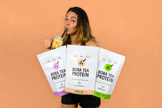 Woman sipping boba tea and holding three Boba Tea Protein bags in classic, matcha, and taro flavors.