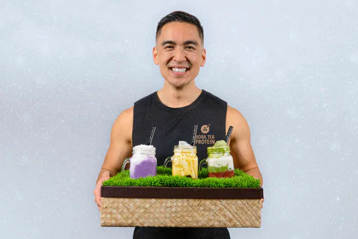 Man holding a tray with four colorful boba drinks in mason jars, wearing a "Boba Tea Protein" tank top.