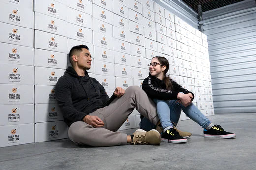 Two people sitting on the floor smiling at each other in front of stacked Boba Tea Protein boxes.