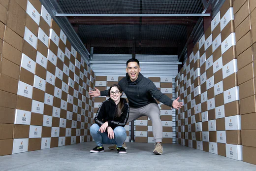 Two people posing in a warehouse aisle surrounded by stacked Boba Tea Protein boxes.