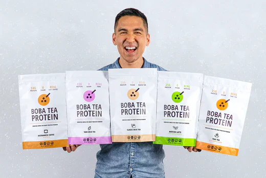 Smiling man holding five Boba Tea Protein bags in various flavors against a light speckled wall.