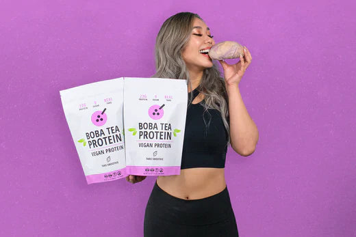 Smiling woman in black activewear holding two Boba Tea Protein vegan bags and eating a taro root.