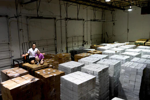 Two people sitting on boxes in a warehouse filled with large stacks of white and brown shipping boxes.