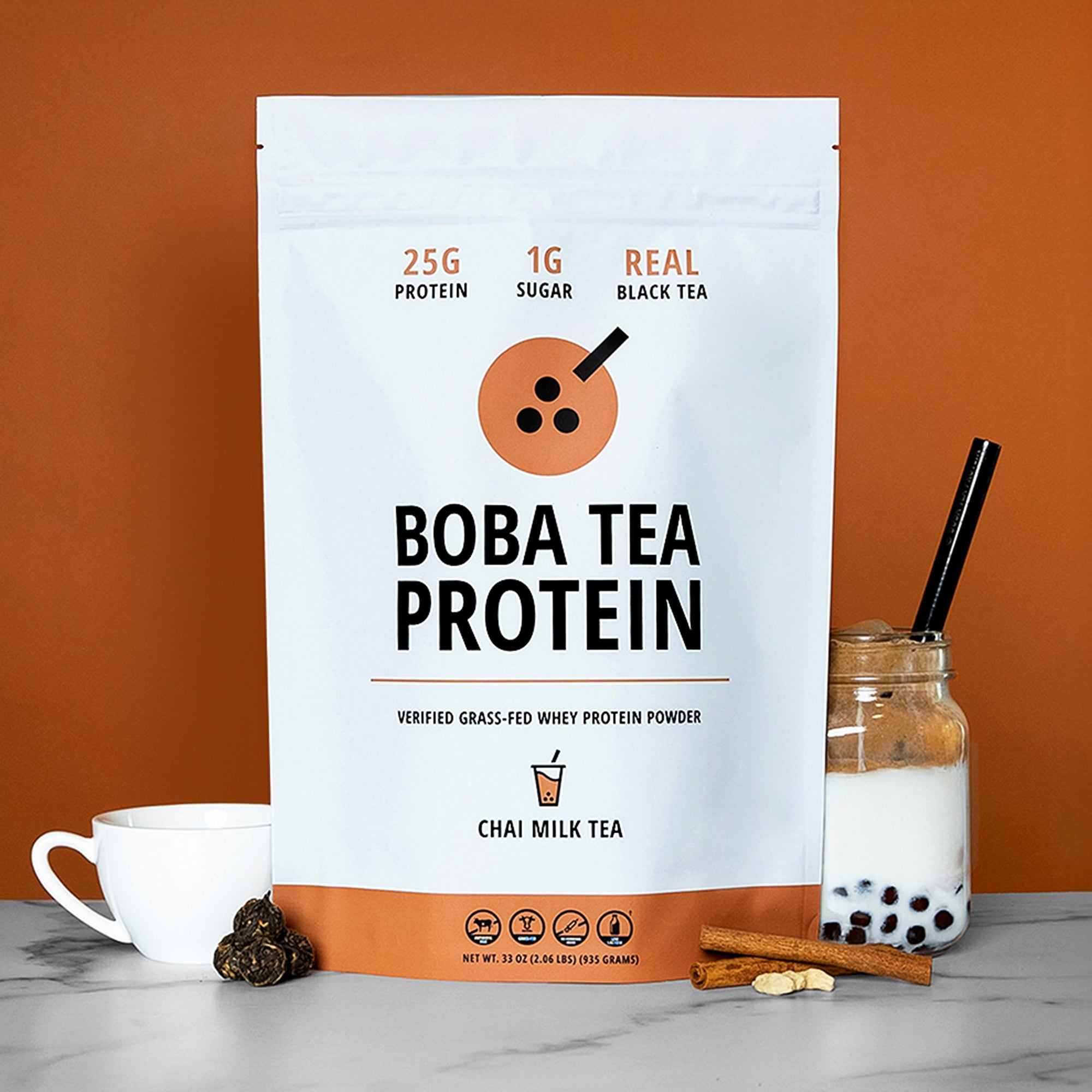 The image shows a bag of "Boba Tea Protein" powder in Chai Milk Tea flavor, featuring highlights like "25g Protein", "1g Sugar", and "Real Black Tea", against a background of light brown matching the chai theme. In front are tea leaves, cinnamon sticks and cardamom seeds, a teacup, and a jar of Boba Tea Protein's Chai Milk Tea poured into milk with tapioca pearls and a metal straw.