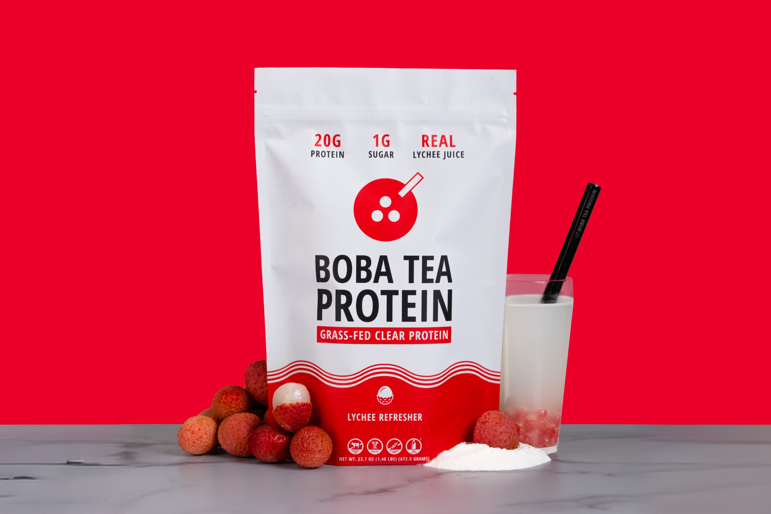 A stand-up pouch of Boba Tea Protein grass-fed clear protein powder, shown with a glass of white drink and lychees on a marble surface against a bright red background.