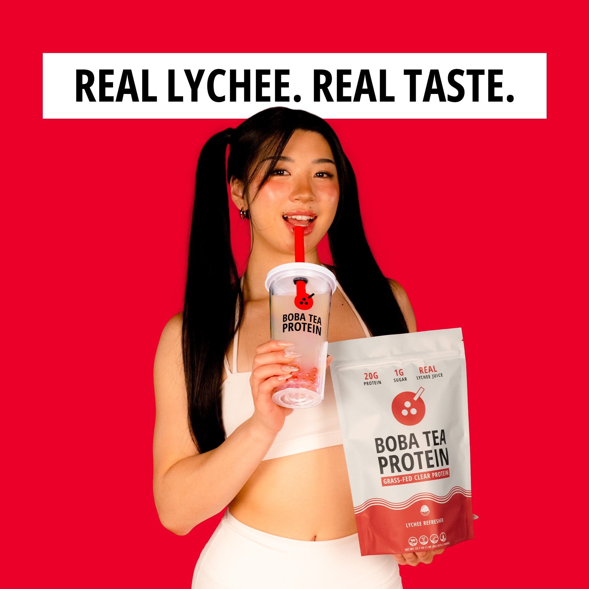 A woman in a white sports bra and leggings holds a clear plastic cup with a red straw and a bag of Boba Tea Protein powder against a solid red background. A white banner at the top reads 'REAL LYCHEE.